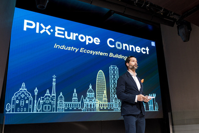 Man standing on a stage speaking to an audience with the event title and logo on a screen behind him